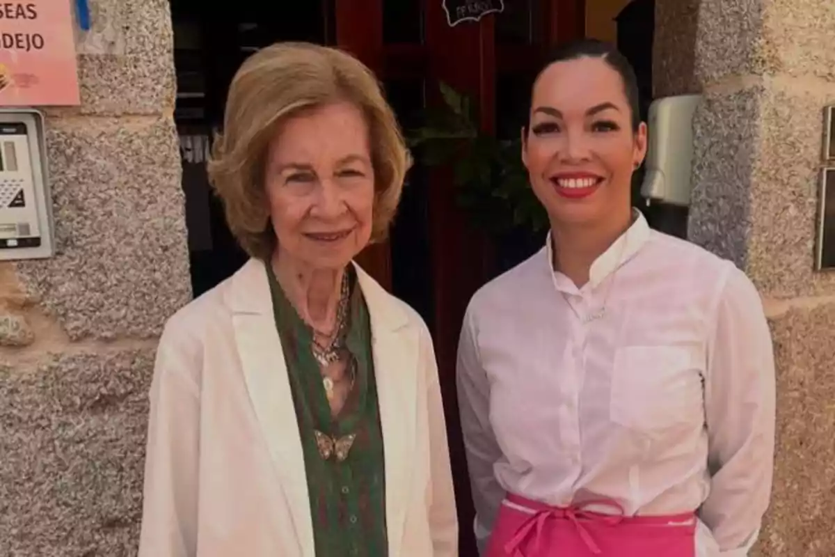 Two women pose smiling in front of the entrance to a stone building; one is wearing a white coat and the other is dressed in a white jersey with a pink apron. Two women pose smiling in front of the entrance to a stone building; one is wearing a white coat and the other is dressed in a white jersey with a pink apron.