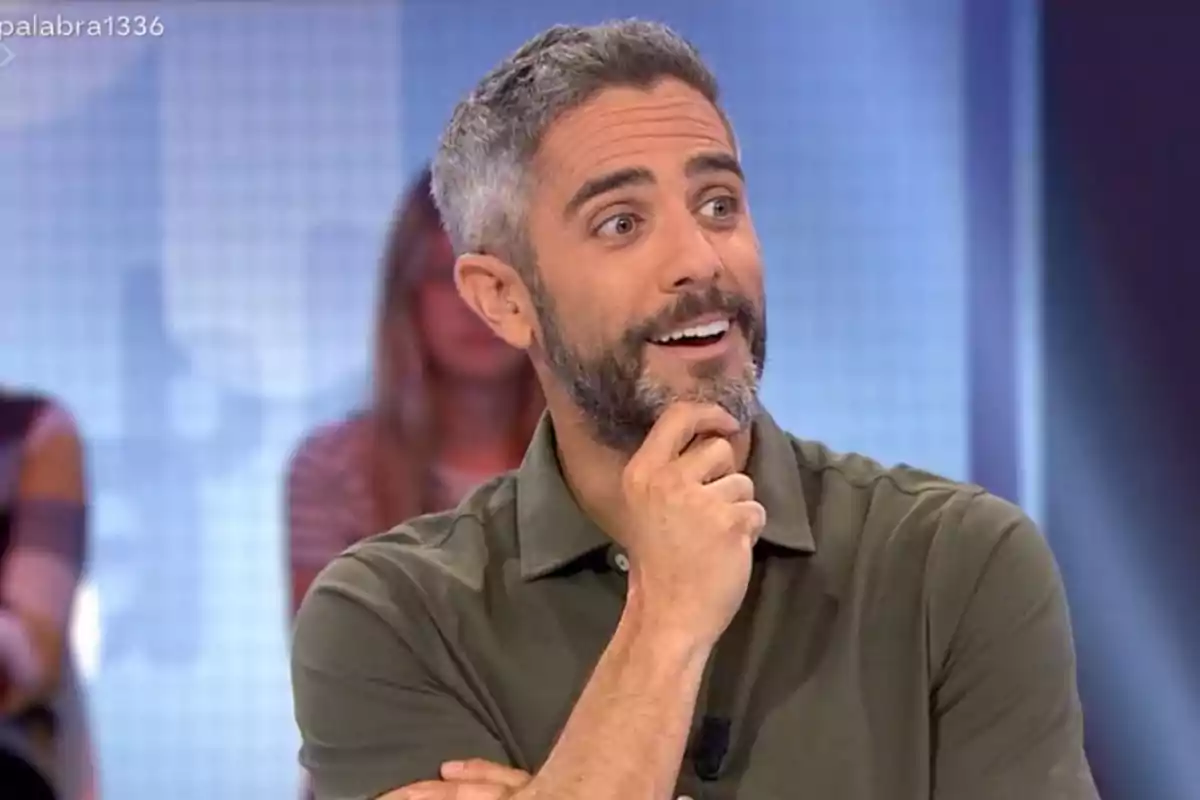 Home de cabell curt i barba amb expressió somrient i pensativa, vestit amb camisa verda, en un estudi de televisió amb persones desenfocades al fons Home de cabell curt i barba amb expressió somrient i pensativa, vestit amb camisa verda, en un estudi de televisió amb persones desenfocades al fons
