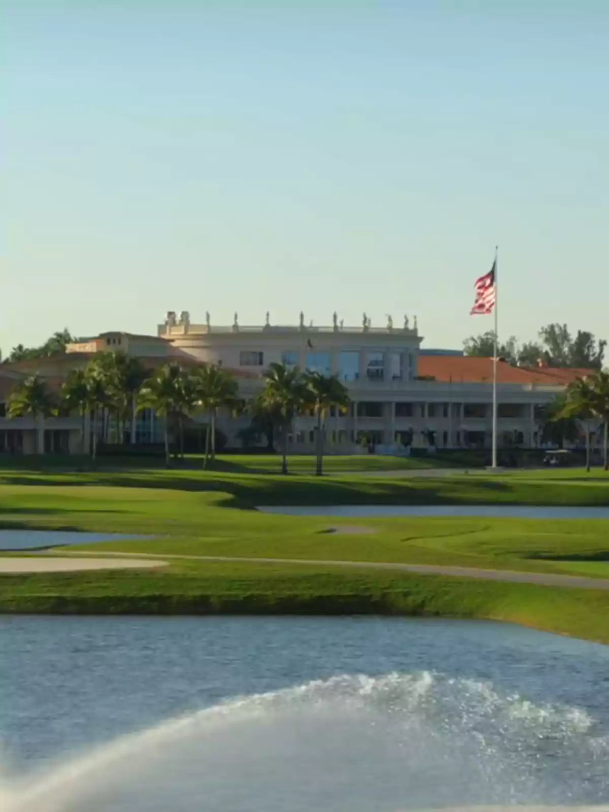 Edificio elegante al fondo rodeado de palmeras con una bandera de Estados Unidos ondeando y un campo de golf con lago y fuente en primer plano