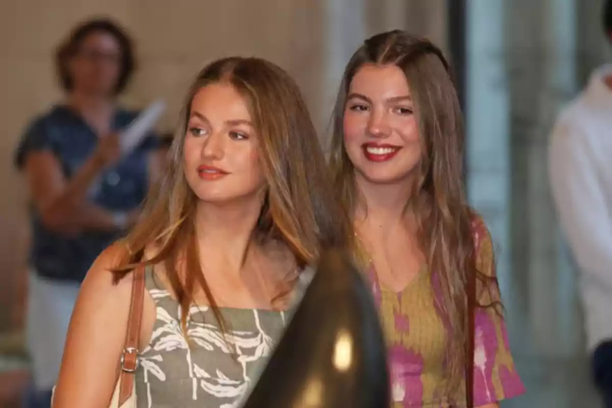 La infanta Sofía y la princesa Leonor jóvenes con el cabello suelto y largo están de pie una al lado de la otra en un ambiente interior, ambas lucen vestidos veraniegos y sonríen mientras miran en diferentes direcciones La infanta Sofía y la princesa Leonor jóvenes con el cabello suelto y largo están de pie una al lado de la otra en un ambiente interior, ambas lucen vestidos veraniegos y sonríen mientras miran en diferentes direcciones
