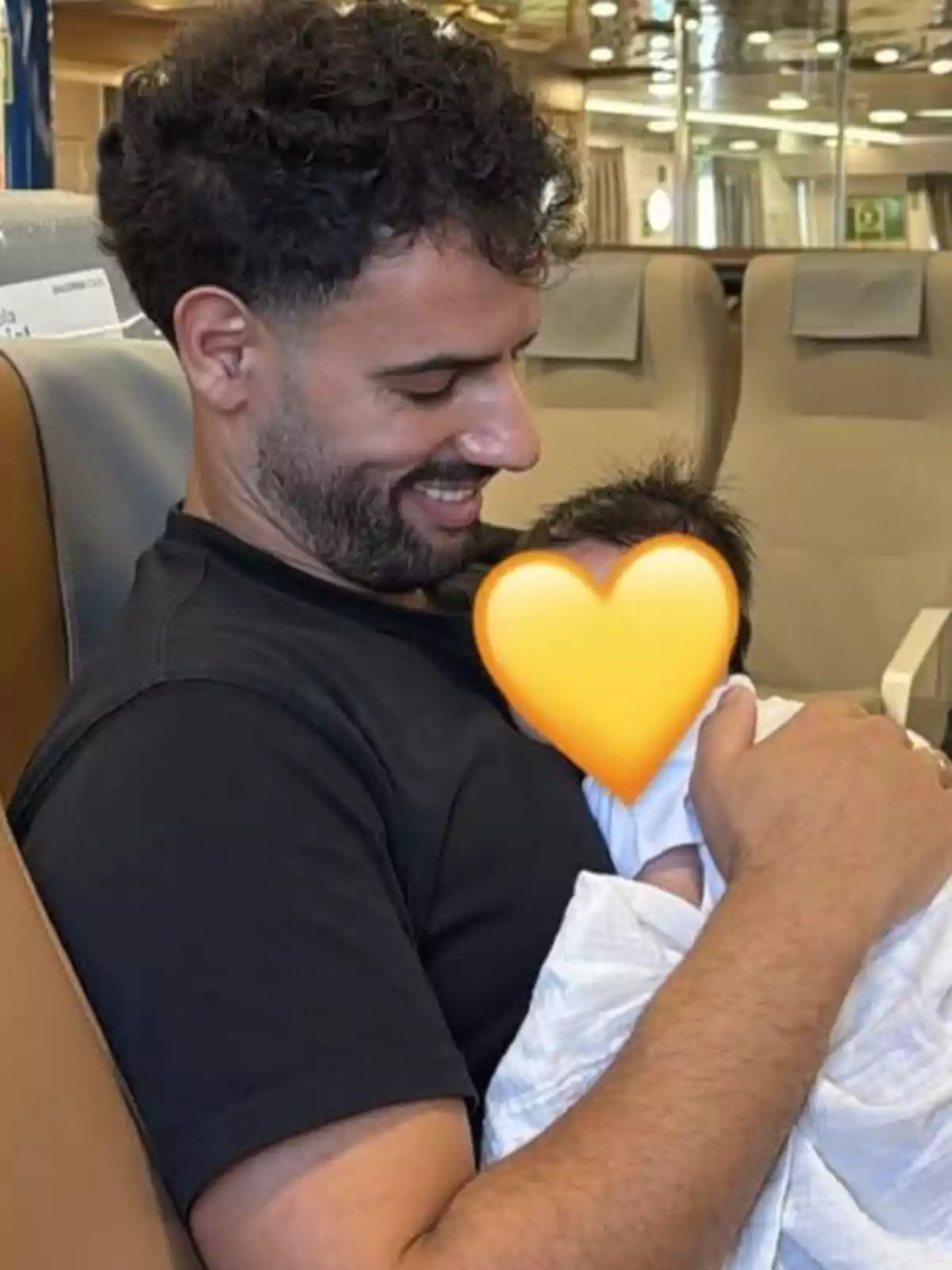 Home somrient sosté en braços un nadó embolcallat amb una manta blanca amb un emoji de cor groc cobrint la cara del nadó Home somrient sosté en braços un nadó embolcallat amb una manta blanca amb un emoji de cor groc cobrint la cara del nadó