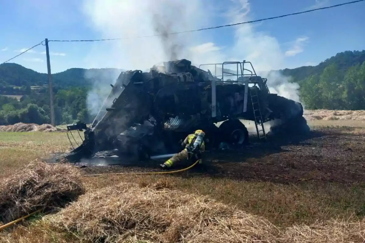 Un bomber apaga l’incendi d’una recol·lectora calcinada en un camp envoltat de muntanyes i bales de fenc.