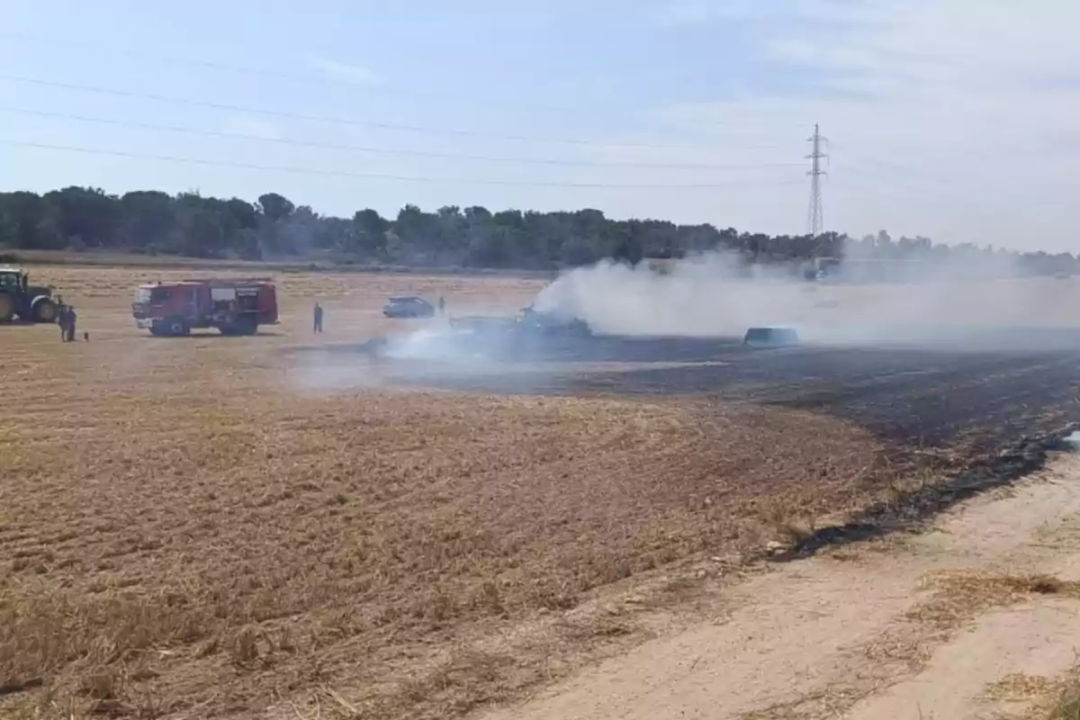 Un campo de cultivo parcialmente quemado con humo saliendo de la zona afectada mientras un camión de bomberos, un tractor y varias personas trabajan en el lugar