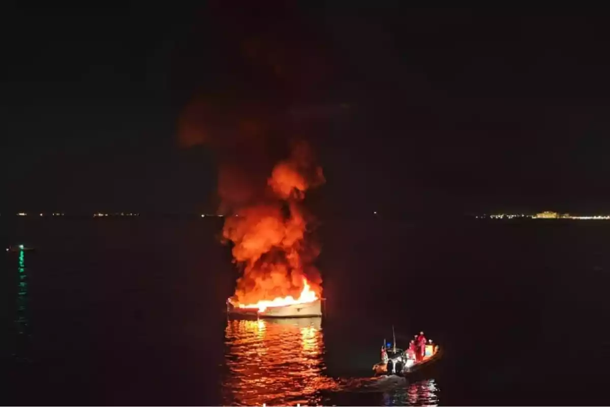 Barco en llamas en el mar durante la noche mientras una lancha de rescate se acerca