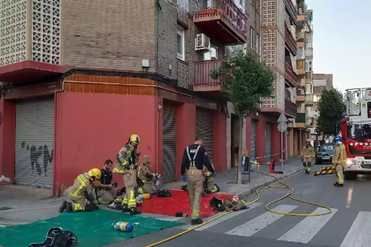 Diversos bombers amb equip de protecció treballen a la vorera davant d’un edifici d’apartaments mentre un camió de bombers està estacionat al carrer.