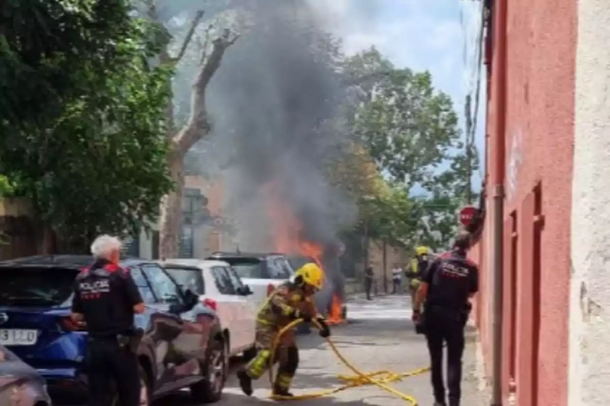 Un bomber amb mànega s’acosta a un cotxe en flames mentre dos policies observen en un carrer urbà.