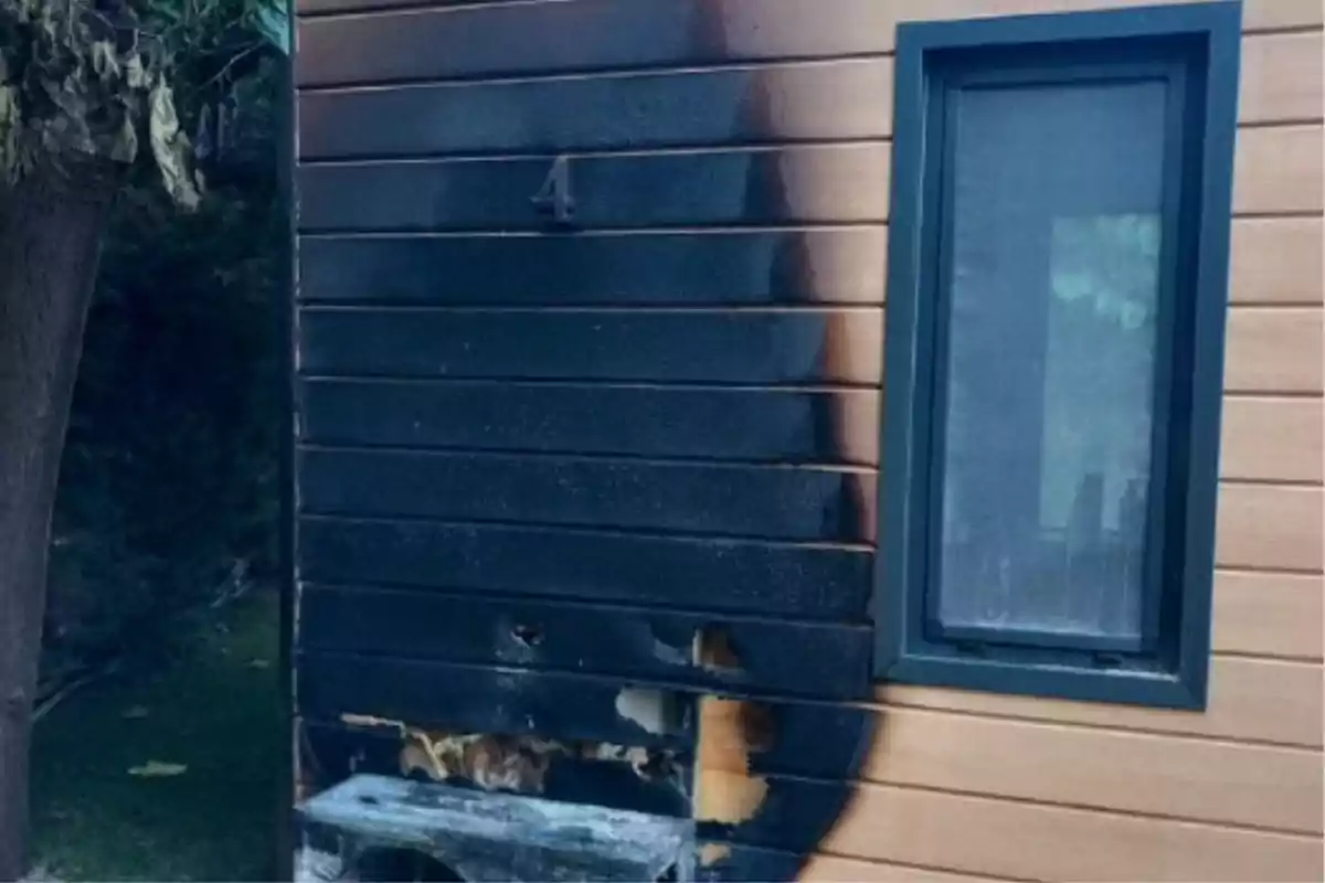 Pared de madera de una casa con una gran mancha negra de quemadura alrededor de una ventana y daños visibles en la parte inferior