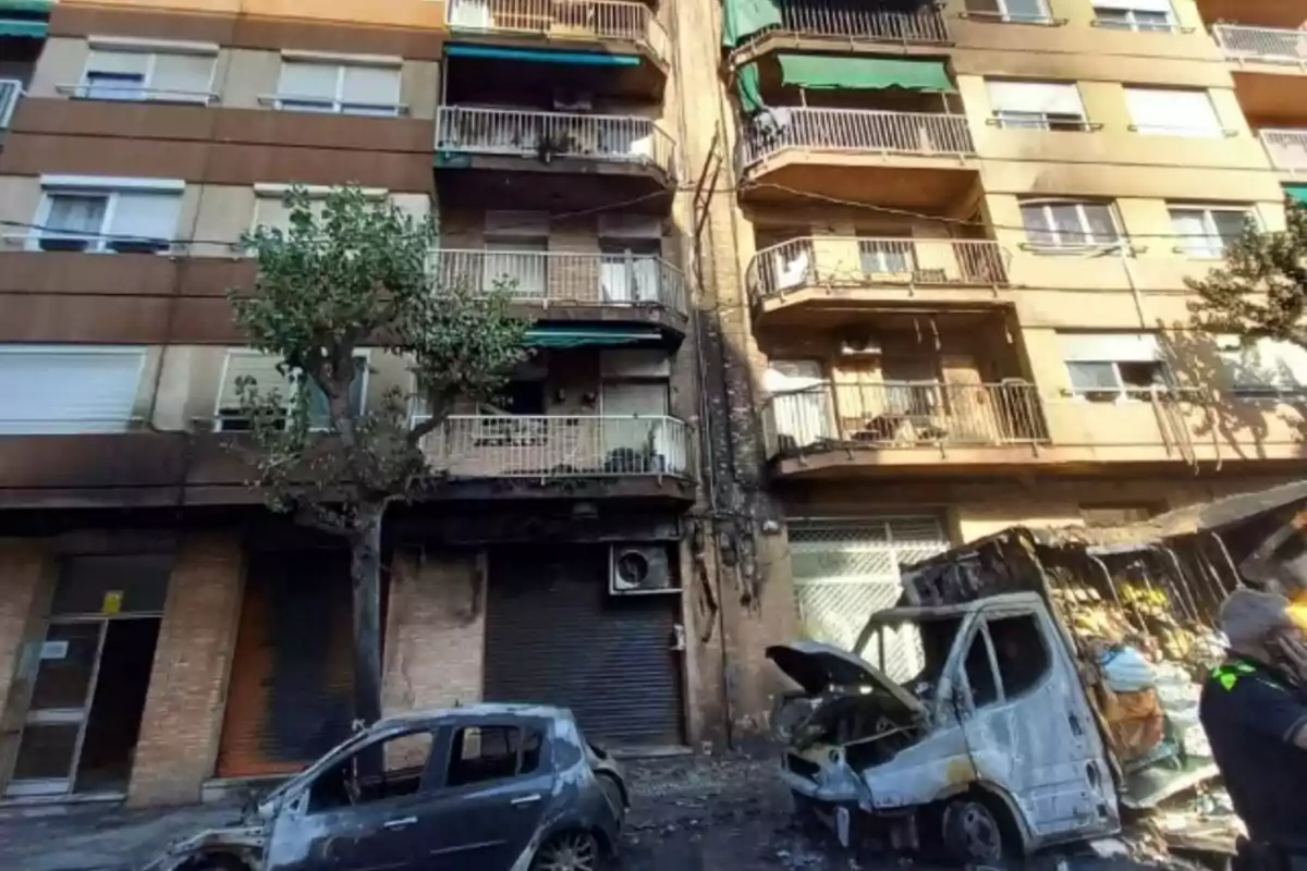 Façana d’edifici amb danys visibles per incendi i dos vehicles calcinats al carrer davant seu