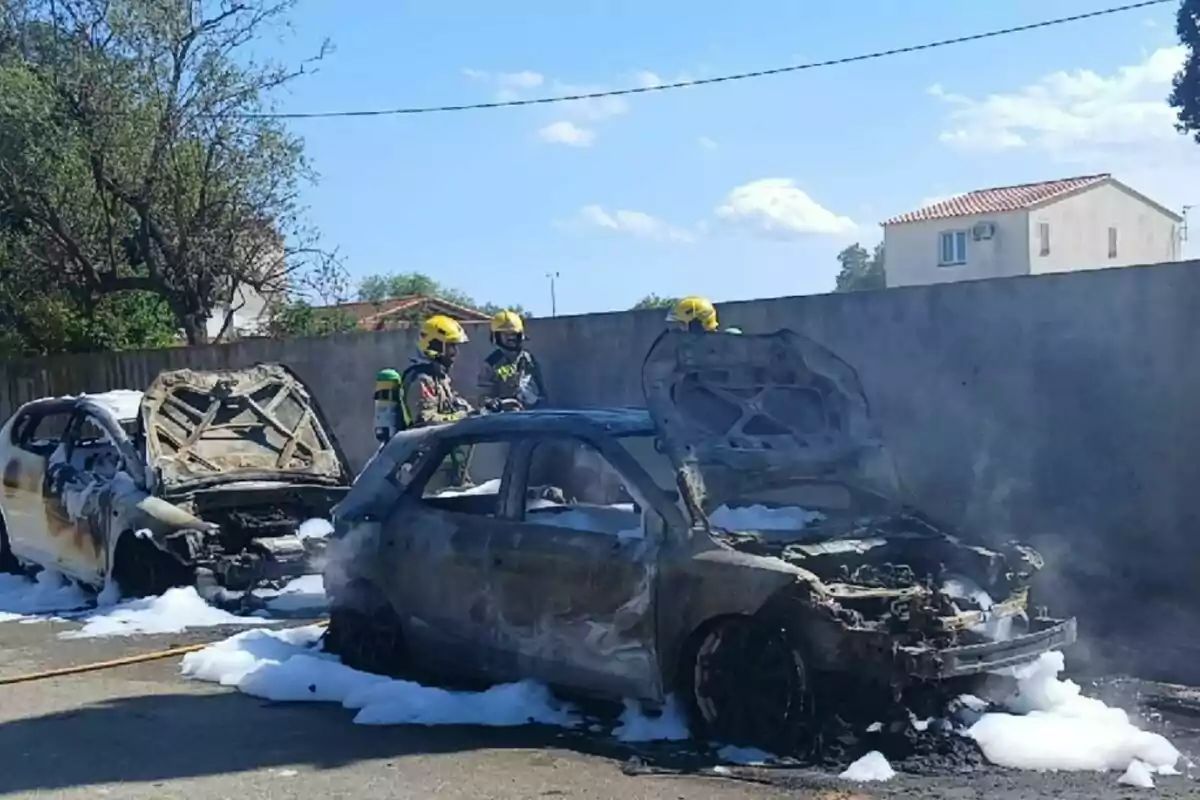 Dos cotxes completament calcinats amb escuma al voltant i tres bombers amb casc groc revisant l’escena en un carrer al costat d’una paret