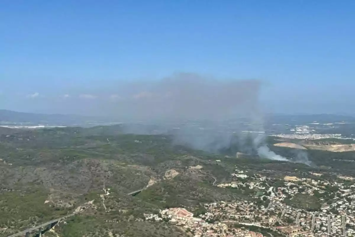 Vista aèria d’una zona boscosa amb fum que s’eleva a causa d’un incendi forestal a prop d’una zona urbana sota un cel serè