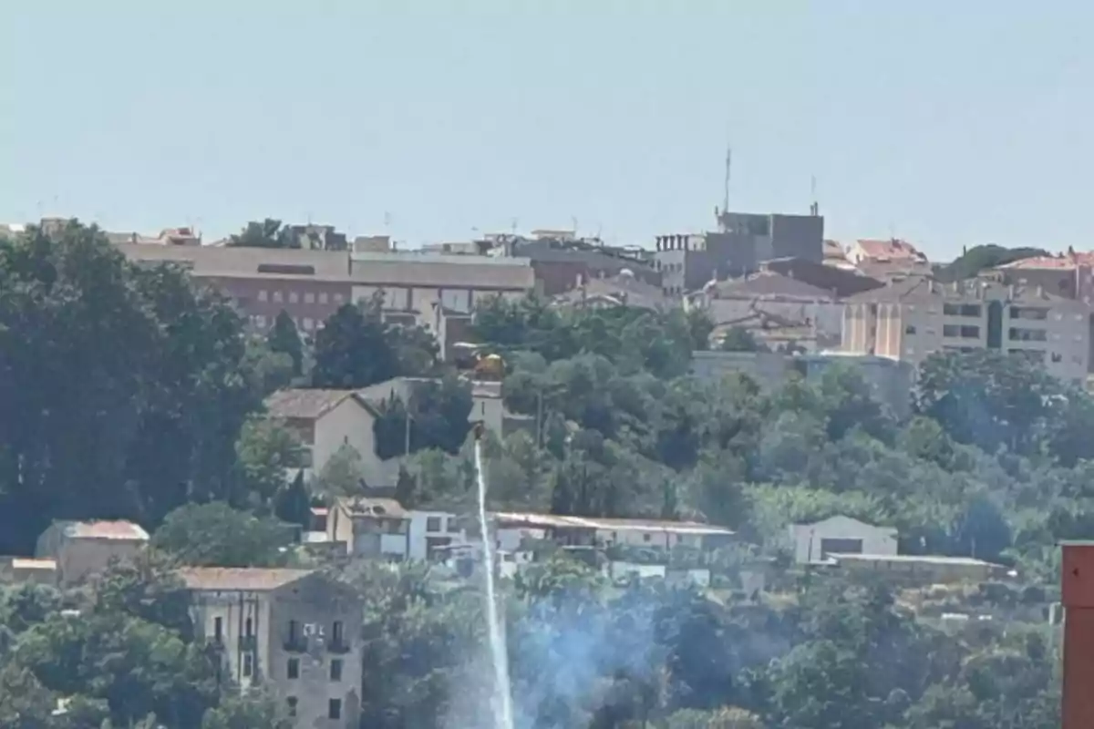 Un helicóptero arroja agua sobre una zona con humo cerca de edificios y vegetación en una ciudad.