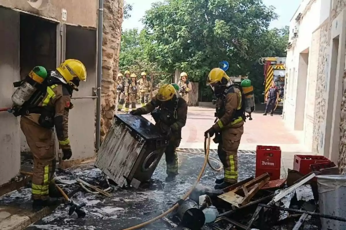 Bombers amb equip de protecció apaguen un incendi en una rentadora cremada i altres objectes al carrer davant d’una casa mentre altres bombers observen al fons.
