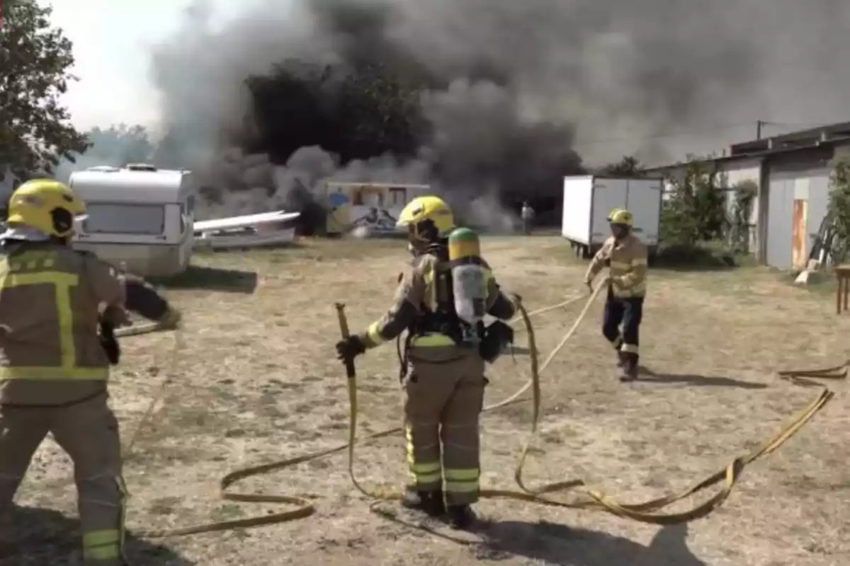 Bomberos con equipo de protección y mangueras trabajan para controlar un incendio en una zona al aire libre con humo negro denso al fondo