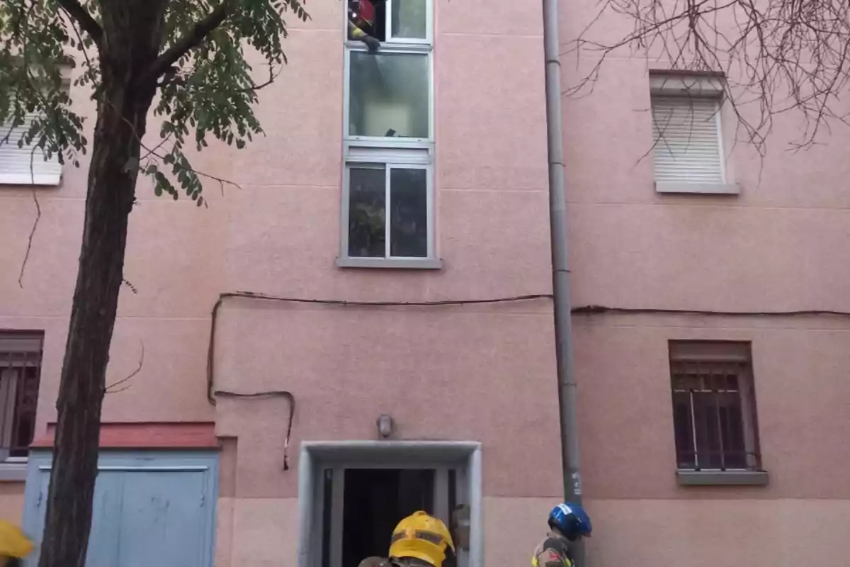 Bomberos con casco amarillo y azul trabajan en el rescate de una persona desde la ventana de un edificio de color rosa