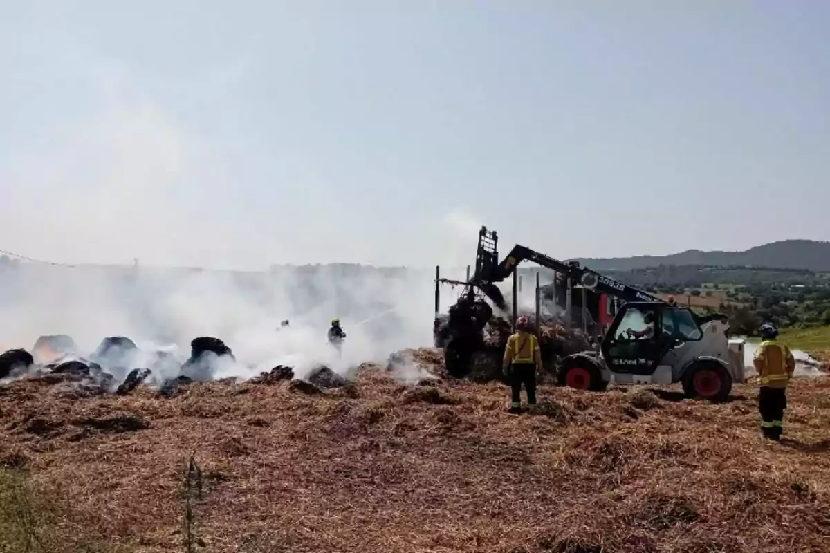 Bombers treballen per apagar un incendi en un camp de bales de palla fent servir mànegues i maquinària pesada mentre el fum s’eleva en l’ambient rural.