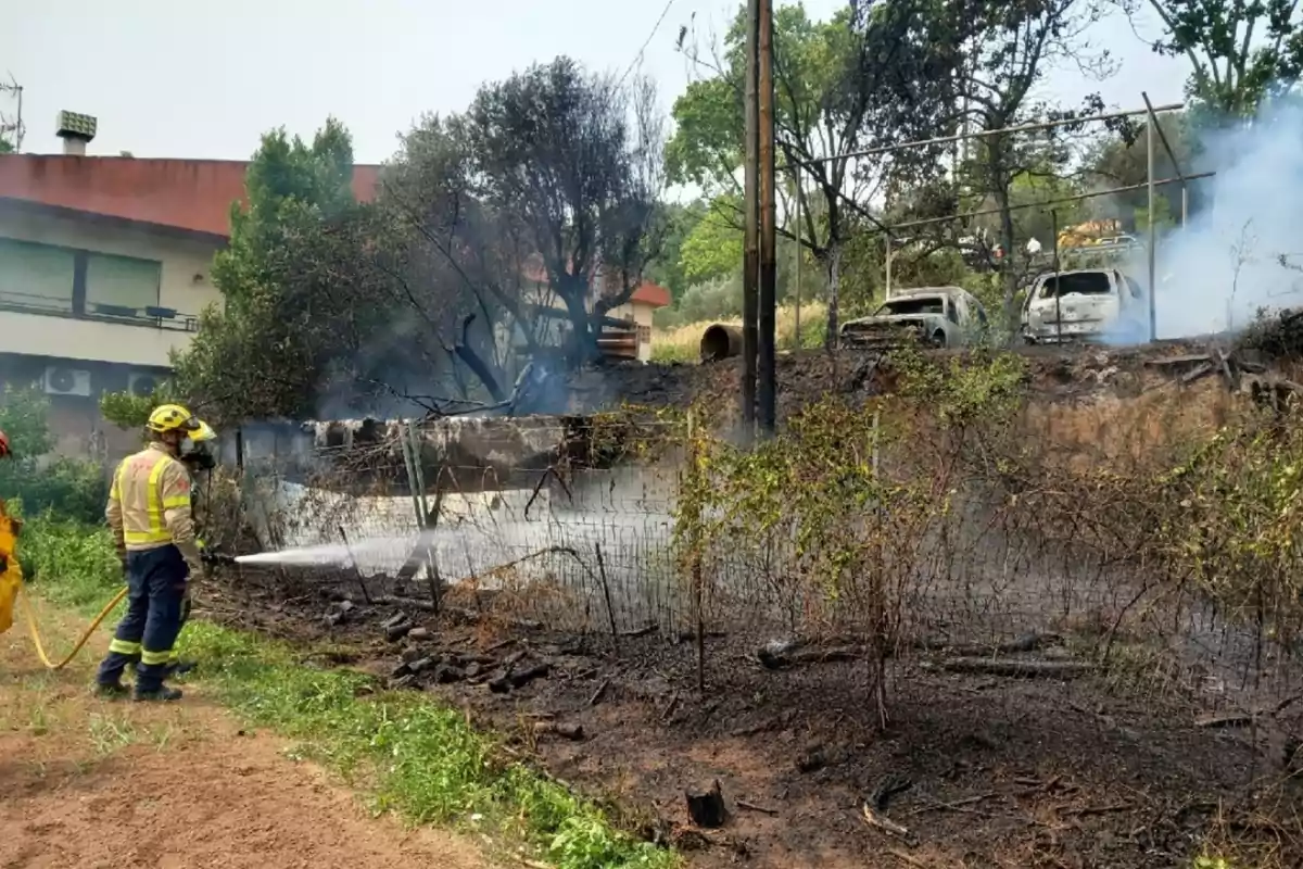 Bombero apaga un incendio en un terreno con vegetación quemada y dos autos calcinados mientras sale humo en el fondo