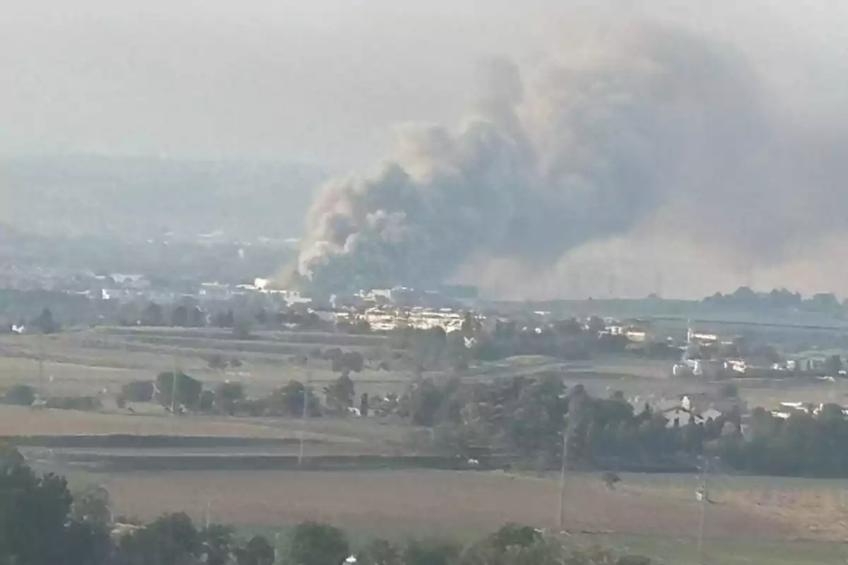 Se observa una gran columna de humo gris elevándose desde una zona urbana rodeada de campos y vegetación