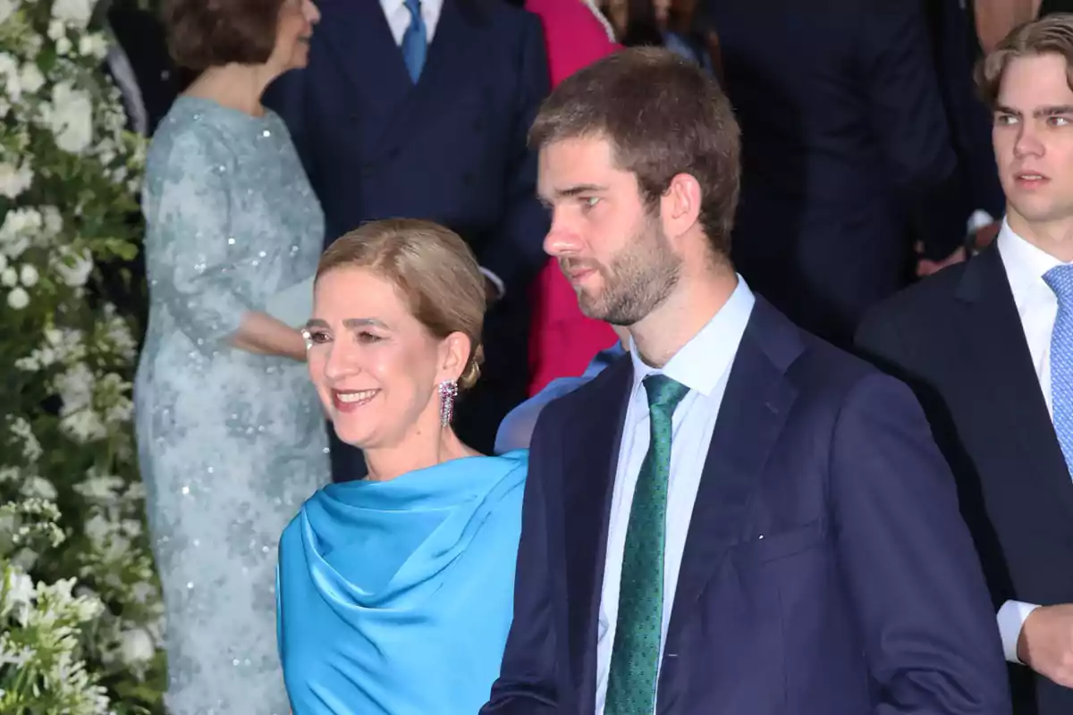 La infanta Cristina con vestido azul y Juan Urdangarin con traje oscuro caminan juntos en un evento elegante rodeados de otras personas vestidas de manera formal y flores blancas al fondo La infanta Cristina con vestido azul y Juan Urdangarin con traje oscuro caminan juntos en un evento elegante rodeados de otras personas vestidas de manera formal y flores blancas al fondo