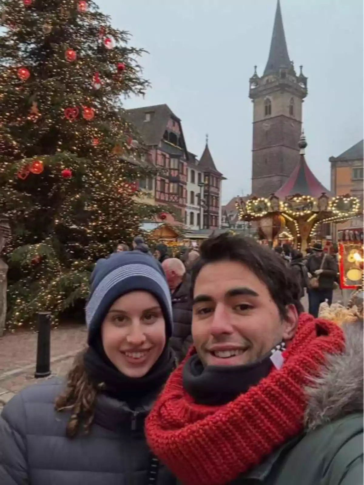 Parella jove somrient davant d’un arbre de Nadal decorat i un mercat nadalenc en una plaça europea amb edificis antics i un campanar al fons. Parella jove somrient davant d’un arbre de Nadal decorat i un mercat nadalenc en una plaça europea amb edificis antics i un campanar al fons.