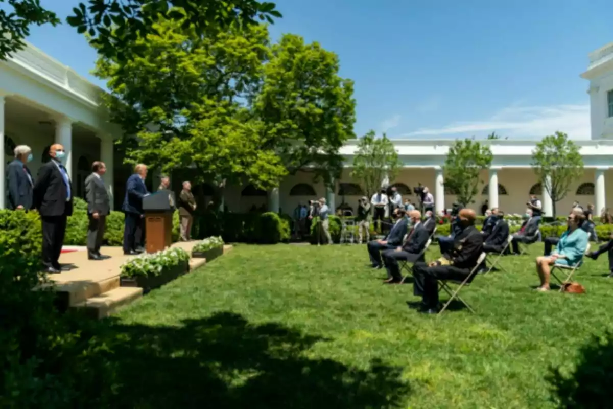 People seated outdoors in a garden listen to a speaker at a podium while others stand near the podium and some attendees wear masks. People seated outdoors in a garden listen to a speaker at a podium while others stand near the podium and some attendees wear masks.