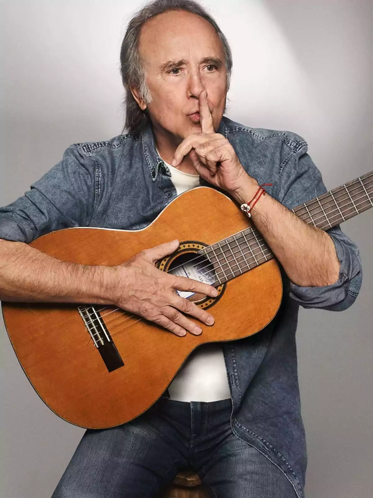 Home de cabells canosos amb camisa texana sosté una guitarra i es porta un dit als llavis en senyal de silenci Home de cabells canosos amb camisa texana sosté una guitarra i es porta un dit als llavis en senyal de silenci