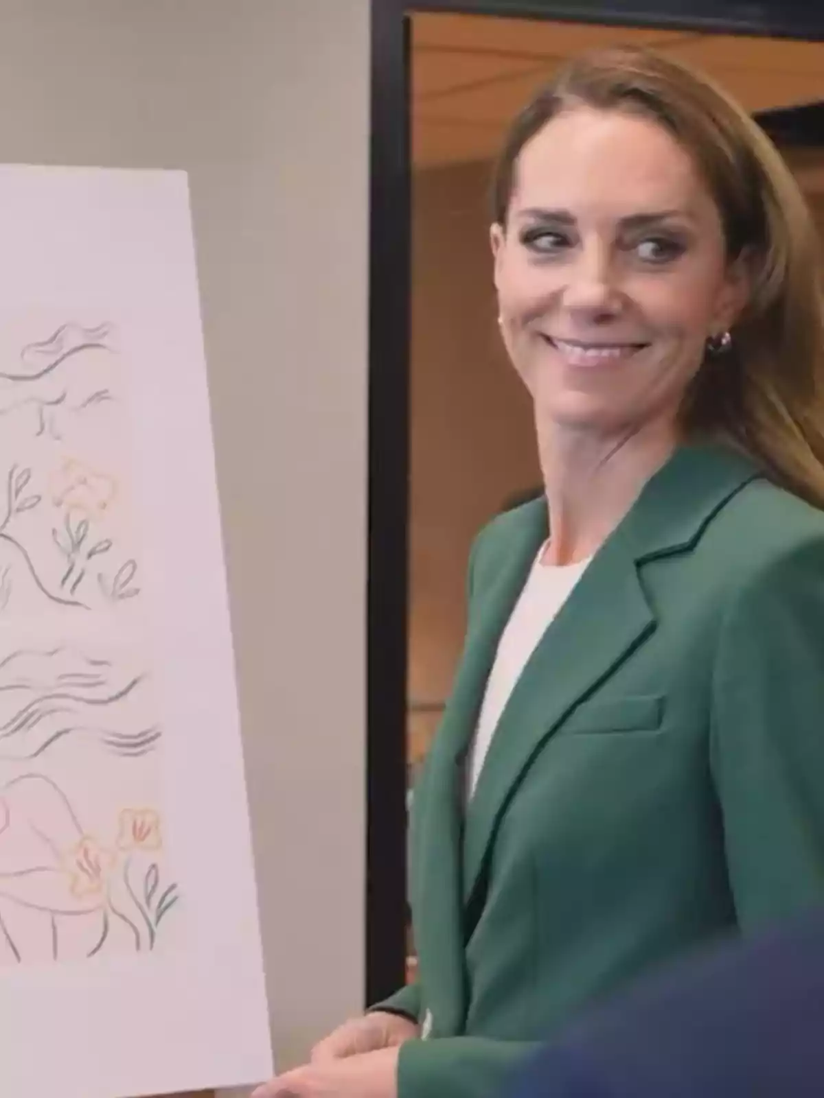 Dona amb jaqueta verda somrient al costat d’una il·lustració en un cavallet Dona amb jaqueta verda somrient al costat d’una il·lustració en un cavallet