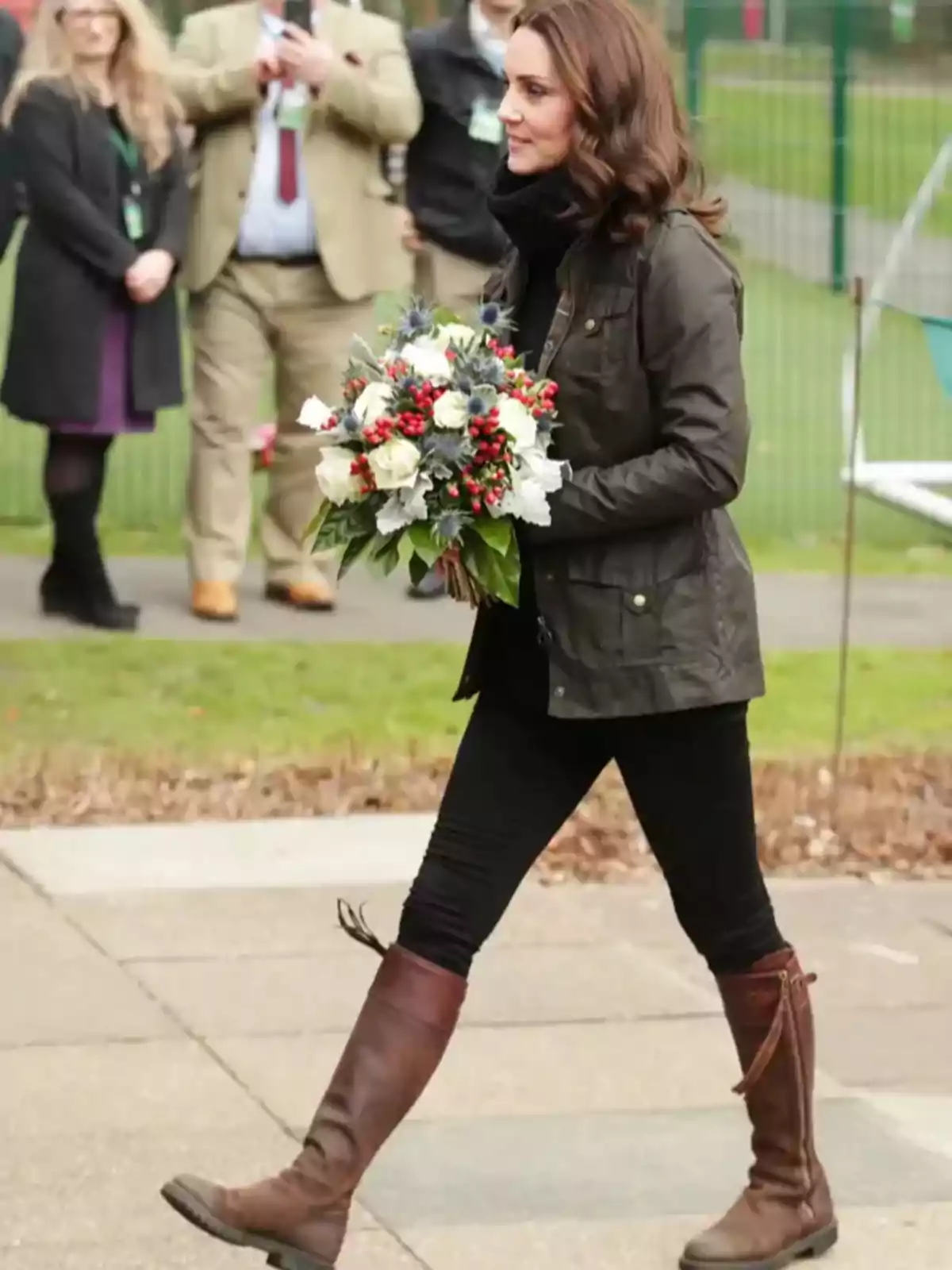 Mujer caminando al aire libre con un ramo de flores en las manos, vestida con chaqueta oscura, pantalones negros y botas marrones, mientras varias personas observan al fondo. Mujer caminando al aire libre con un ramo de flores en las manos, vestida con chaqueta oscura, pantalones negros y botas marrones, mientras varias personas observan al fondo.