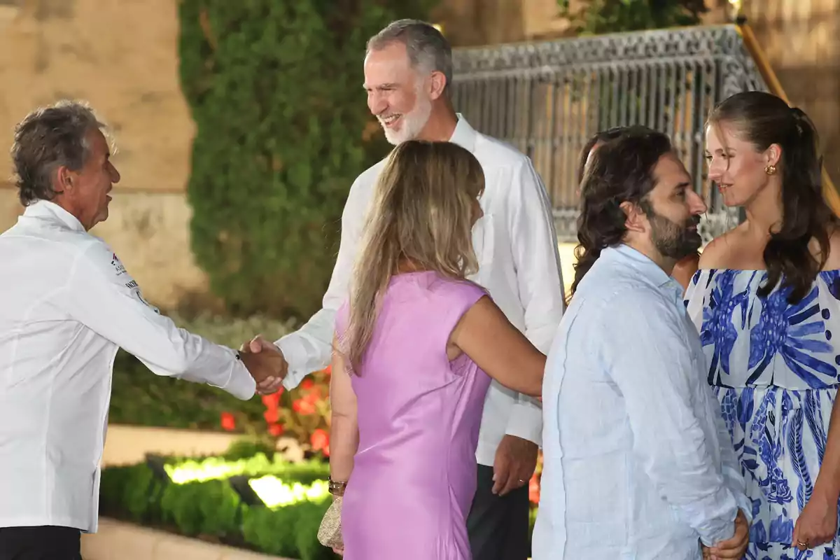 Koldo Royo amb camisa blanca saluda de mà a Felip VI somrient mentre dues parelles conversen al seu costat en un entorn a l’aire lliure amb plantes i flors de fons. Koldo Royo amb camisa blanca saluda de mà a Felip VI somrient mentre dues parelles conversen al seu costat en un entorn a l’aire lliure amb plantes i flors de fons.