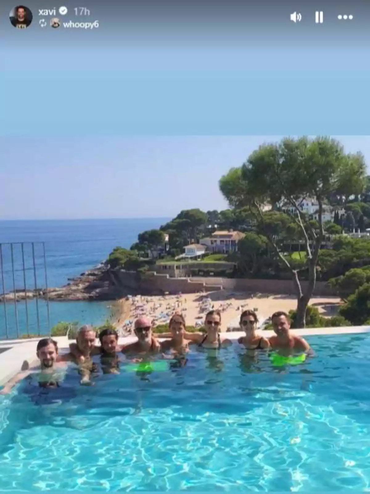 Ocho personas disfrutan en una piscina infinita con vista al mar y una playa al fondo rodeada de árboles y casas Ocho personas disfrutan en una piscina infinita con vista al mar y una playa al fondo rodeada de árboles y casas