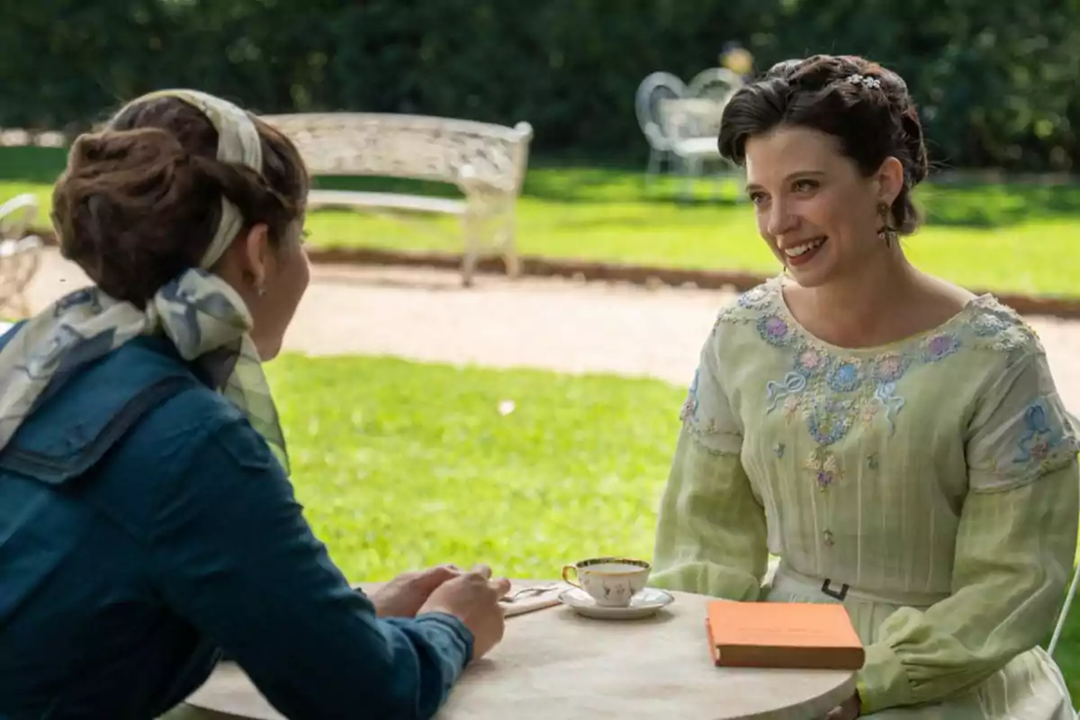 Martina y otra mujer conversan sentadas en una mesa al aire libre en un jardín una de ellas sonríe mientras sostiene una taza de té y la otra tiene un libro naranja sobre la mesa. Captura de 'La Promesa' Martina y otra mujer conversan sentadas en una mesa al aire libre en un jardín una de ellas sonríe mientras sostiene una taza de té y la otra tiene un libro naranja sobre la mesa. Captura de 'La Promesa'
