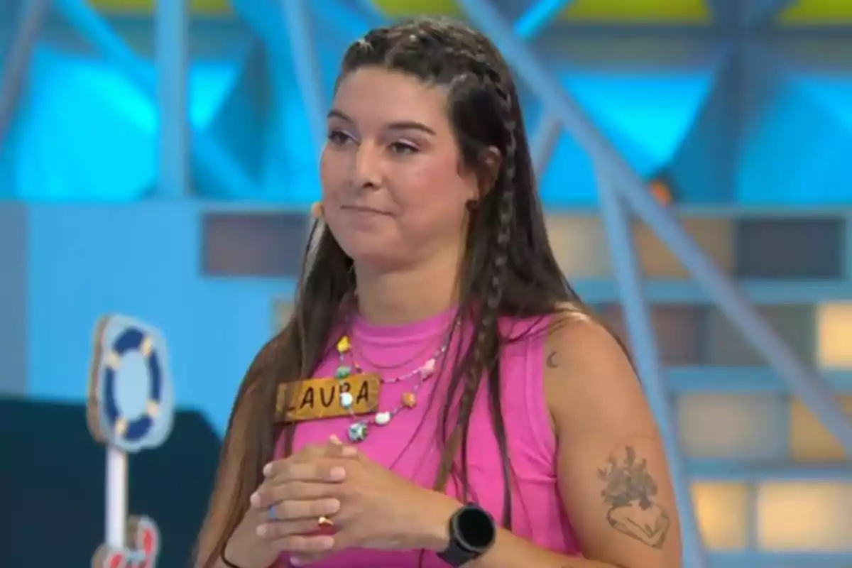 Laura con camiseta rosa y trenzas en el cabello participando en un programa de televisión, lleva un collar colorido, un reloj negro y una placa con el nombre Laura. Captura de 'La Ruleta de la Suerte' Laura con camiseta rosa y trenzas en el cabello participando en un programa de televisión, lleva un collar colorido, un reloj negro y una placa con el nombre Laura. Captura de 'La Ruleta de la Suerte'