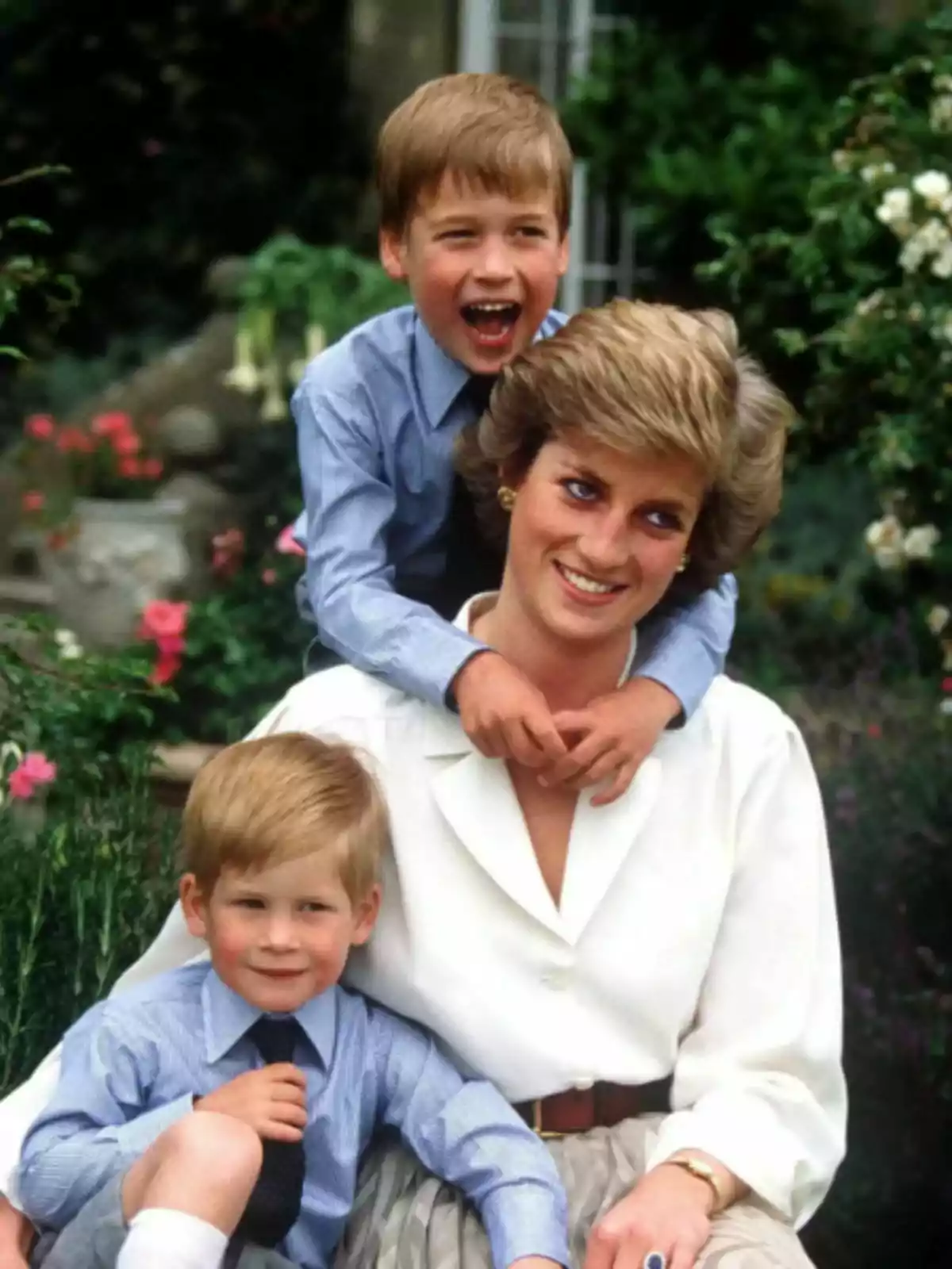 Lady Di somrient asseguda en un jardí amb els prínceps Guillem i Harry abraçant-la, tots vestits de manera elegant i envoltats de flors. Lady Di somrient asseguda en un jardí amb els prínceps Guillem i Harry abraçant-la, tots vestits de manera elegant i envoltats de flors.