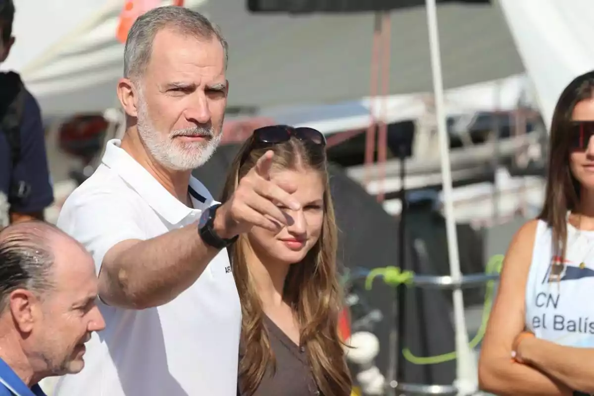 Un hombre de barba canosa señala hacia adelante mientras está acompañado de varias personas en un ambiente al aire libre con barcos de fondo Un hombre de barba canosa señala hacia adelante mientras está acompañado de varias personas en un ambiente al aire libre con barcos de fondo