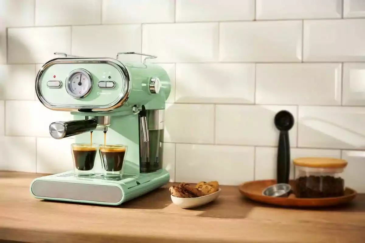Mint green coffee maker brewing two espressos in a kitchen with a white tiled background and a tray with cookies and ground coffee next to it. Mint green coffee maker brewing two espressos in a kitchen with a white tiled background and a tray with cookies and ground coffee next to it.