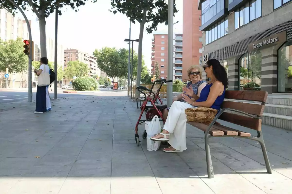Dues dones assegudes en un banc conversen i somriuen en una vorera àmplia i assolellada al costat d’un caminador i bosses de compra mentre una altra persona espera a la cantonada del carrer.