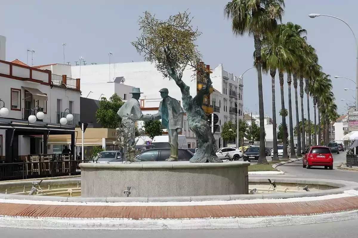 Font en una rotonda amb escultures de dues persones donant-se la mà al costat d’un arbre, envoltada d’edificis, palmeres i cotxes en un carrer urbà. Font en una rotonda amb escultures de dues persones donant-se la mà al costat d’un arbre, envoltada d’edificis, palmeres i cotxes en un carrer urbà.
