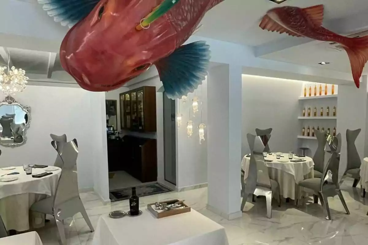 Restaurant elegant amb cadires metàl·liques, taules rodones amb estovalles blanques, ampolles a les prestatgeries i grans figures de peixos penjades del sostre. Restaurant elegant amb cadires metàl·liques, taules rodones amb estovalles blanques, ampolles a les prestatgeries i grans figures de peixos penjades del sostre.