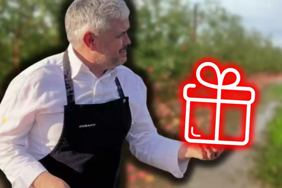 Hombre de cabello canoso con camisa blanca y delantal negro sostiene un dibujo de regalo brillante en un entorno al aire libre con vegetación de fondo