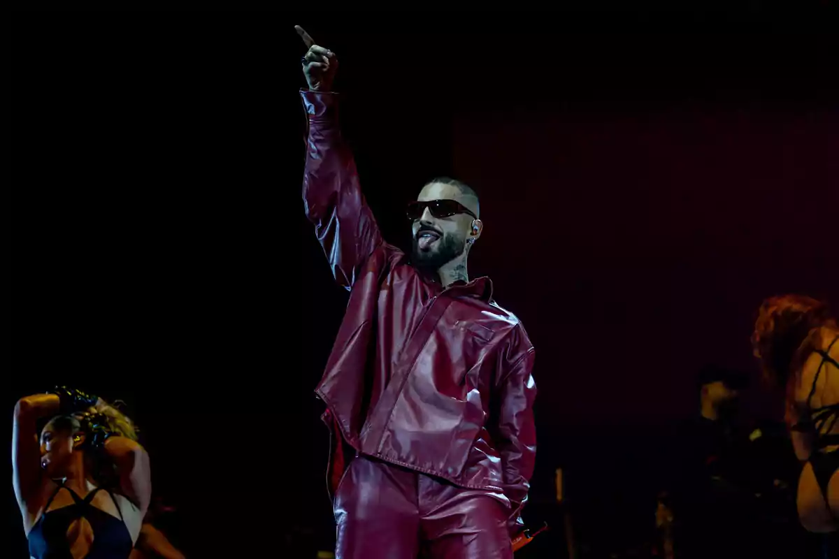Maluma wearing dark sunglasses and a red suit raises one arm while performing on stage accompanied by dancers. Maluma wearing dark sunglasses and a red suit raises one arm while performing on stage accompanied by dancers.