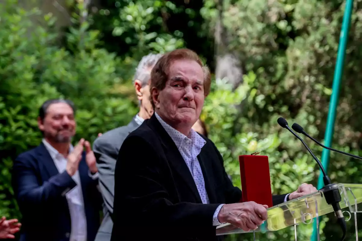 Manuel de la Calva de pie frente a un atril con micrófonos sosteniendo una caja roja mientras varias personas aplauden en el fondo en un entorno al aire libre con vegetación Manuel de la Calva de pie frente a un atril con micrófonos sosteniendo una caja roja mientras varias personas aplauden en el fondo en un entorno al aire libre con vegetación