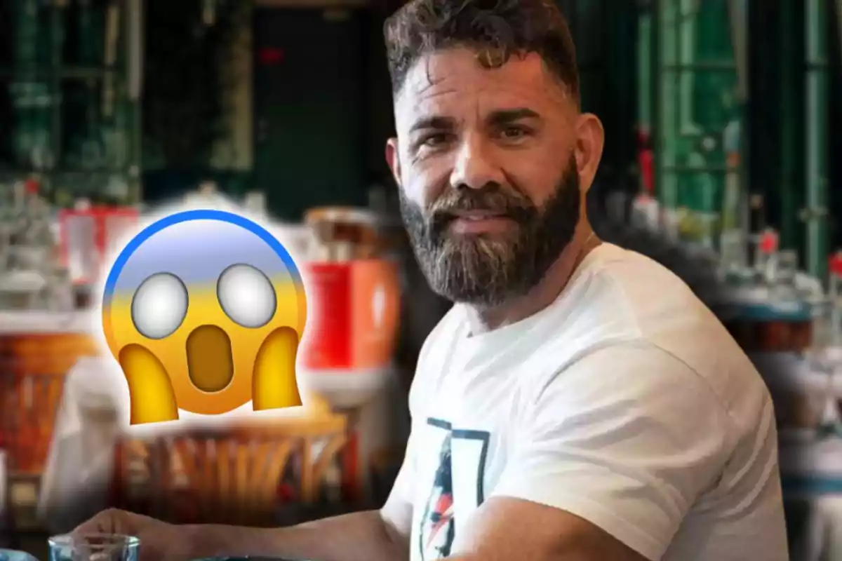 Home amb barba i samarreta blanca assegut en un restaurant amb un emoji de sorpresa al costat