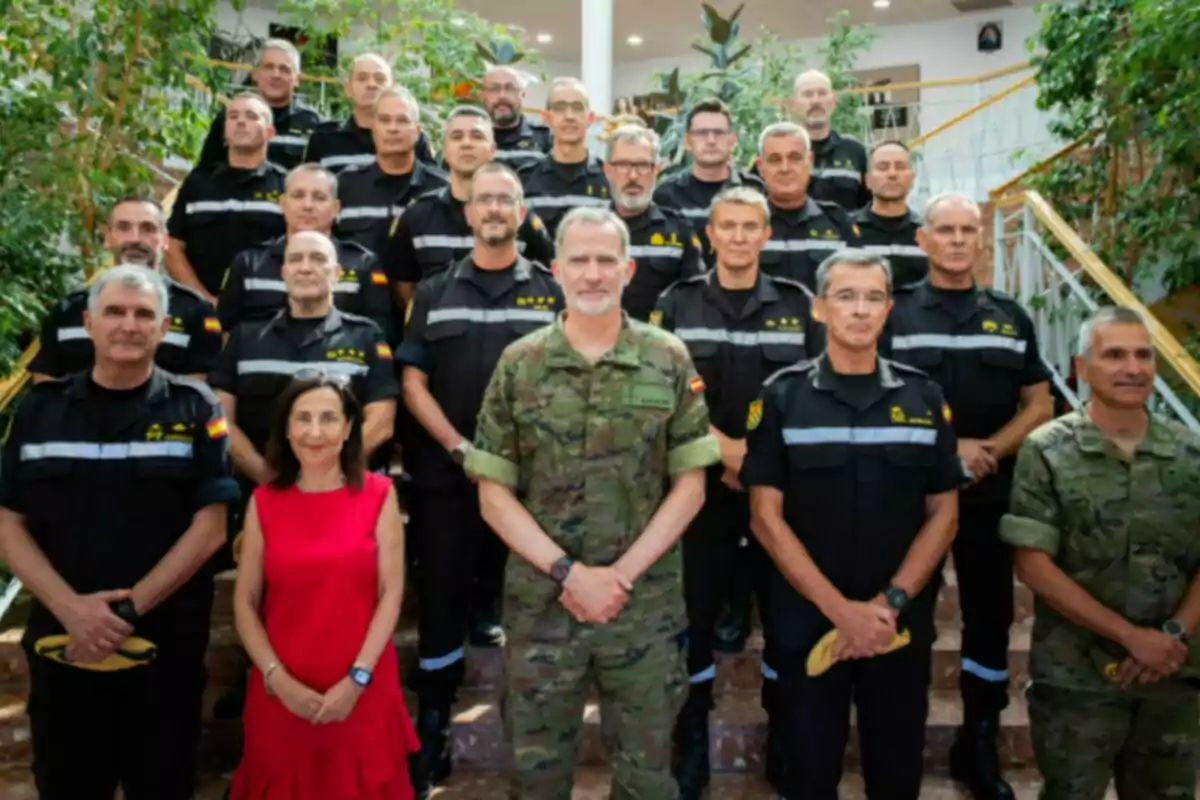 Group of people in military and civil protection uniforms posing on a staircase next to a woman dressed in red in an indoor setting with plants