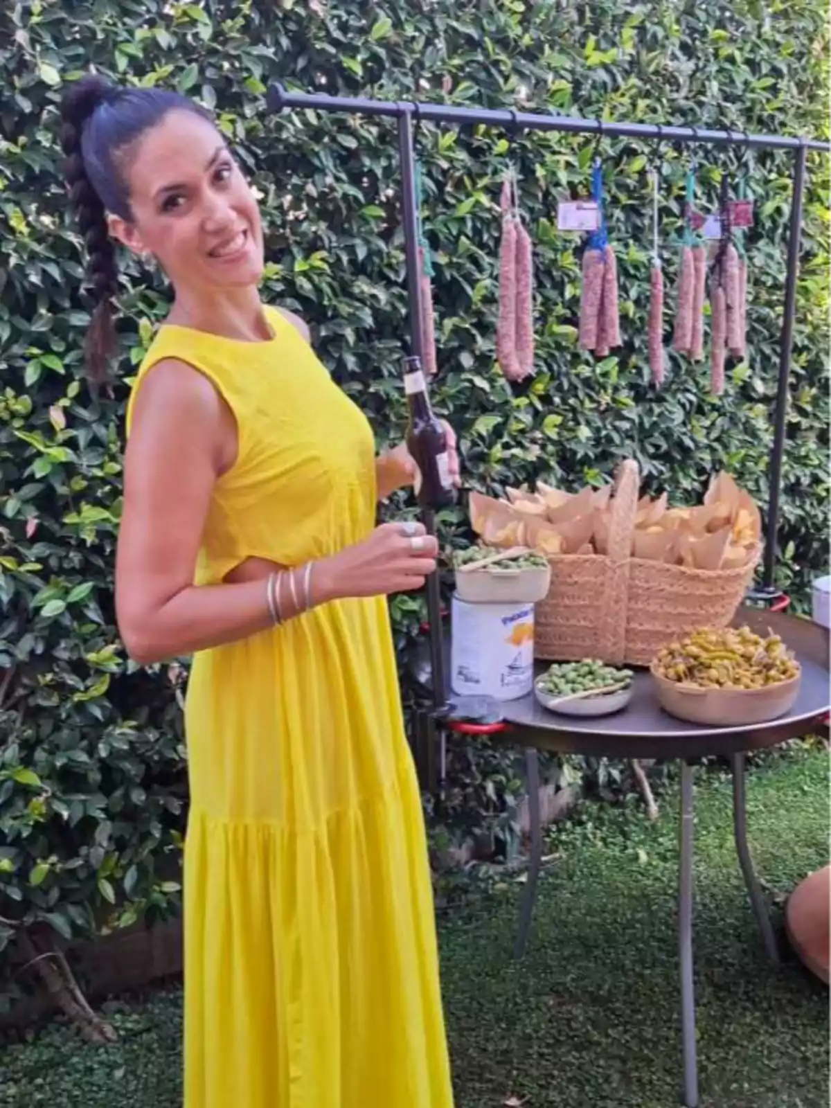 Mujer con vestido amarillo sonríe mientras sostiene una bebida junto a una mesa con aperitivos y embutidos en un jardín Mujer con vestido amarillo sonríe mientras sostiene una bebida junto a una mesa con aperitivos y embutidos en un jardín