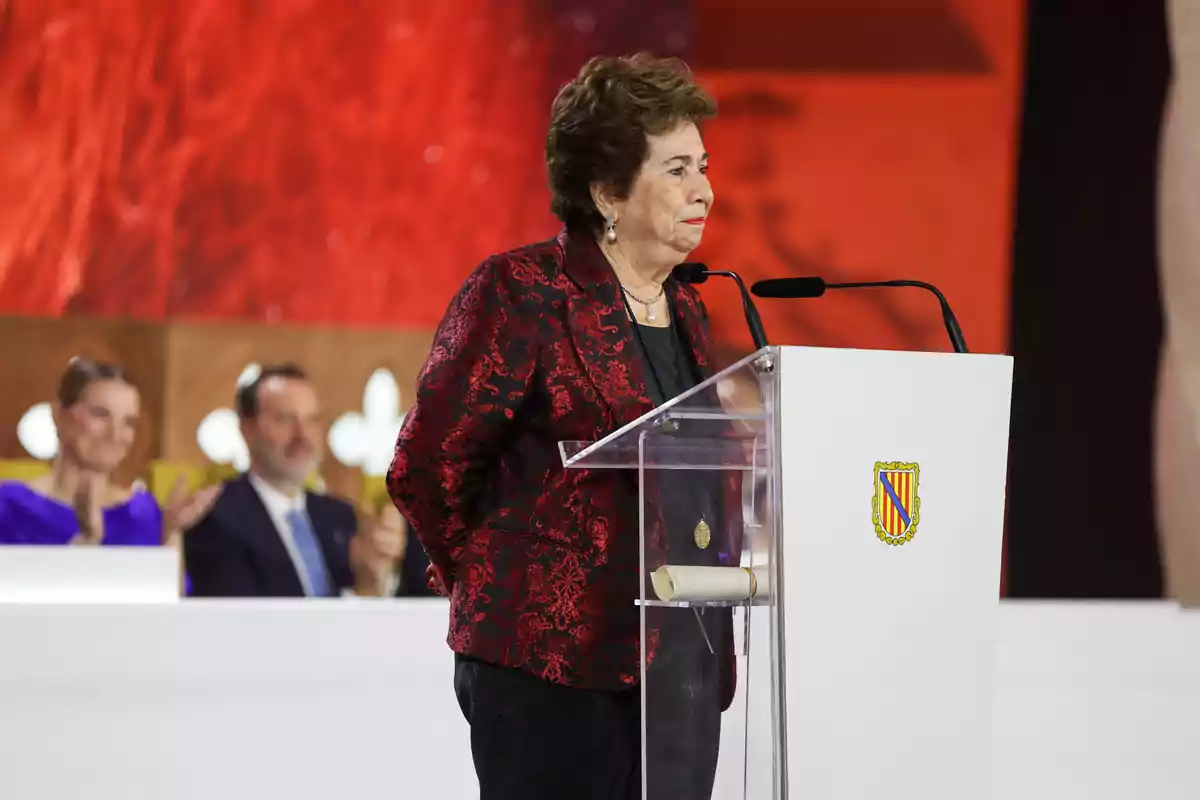 María Luisa Cava de Llano de pie frente a un atril blanco con escudo, usando saco rojo y negro, hablando en un evento formal con fondo rojo y personas aplaudiendo al fondo María Luisa Cava de Llano de pie frente a un atril blanco con escudo, usando saco rojo y negro, hablando en un evento formal con fondo rojo y personas aplaudiendo al fondo
