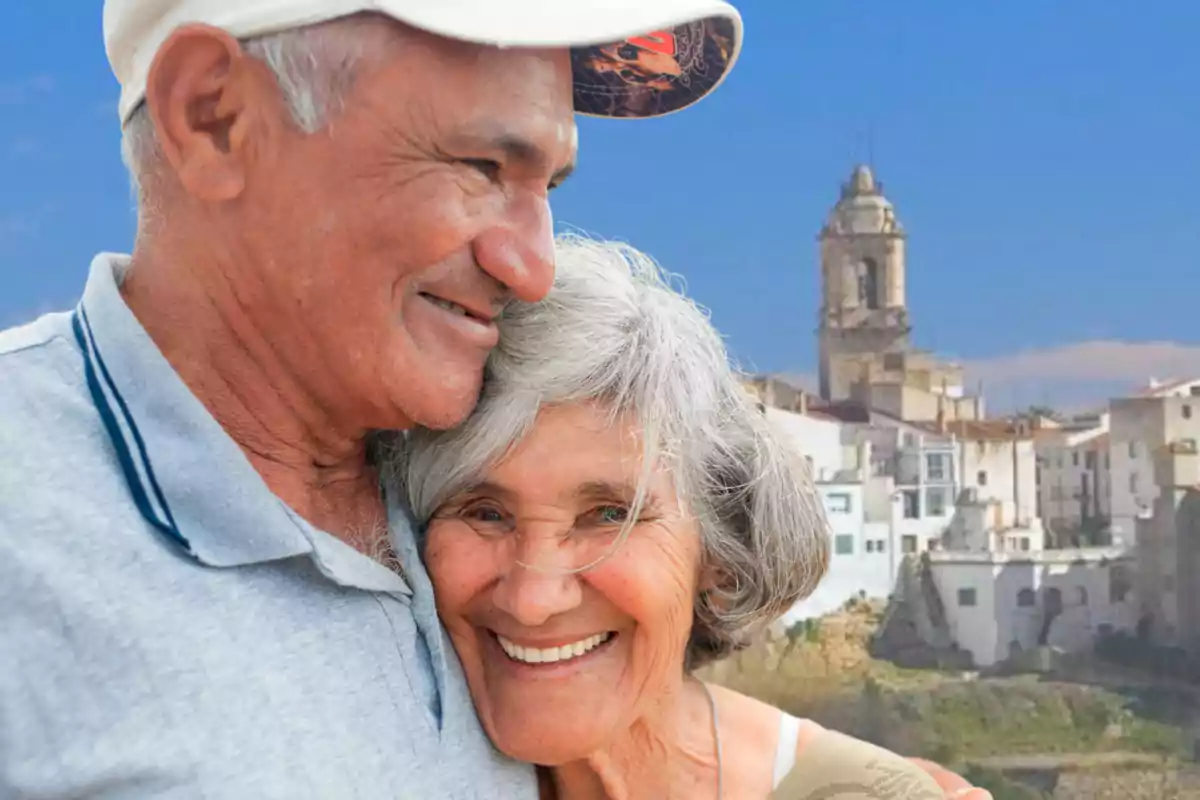Pareja de personas mayores sonriendo y abrazándose con un pueblo y una iglesia al fondo en un día soleado