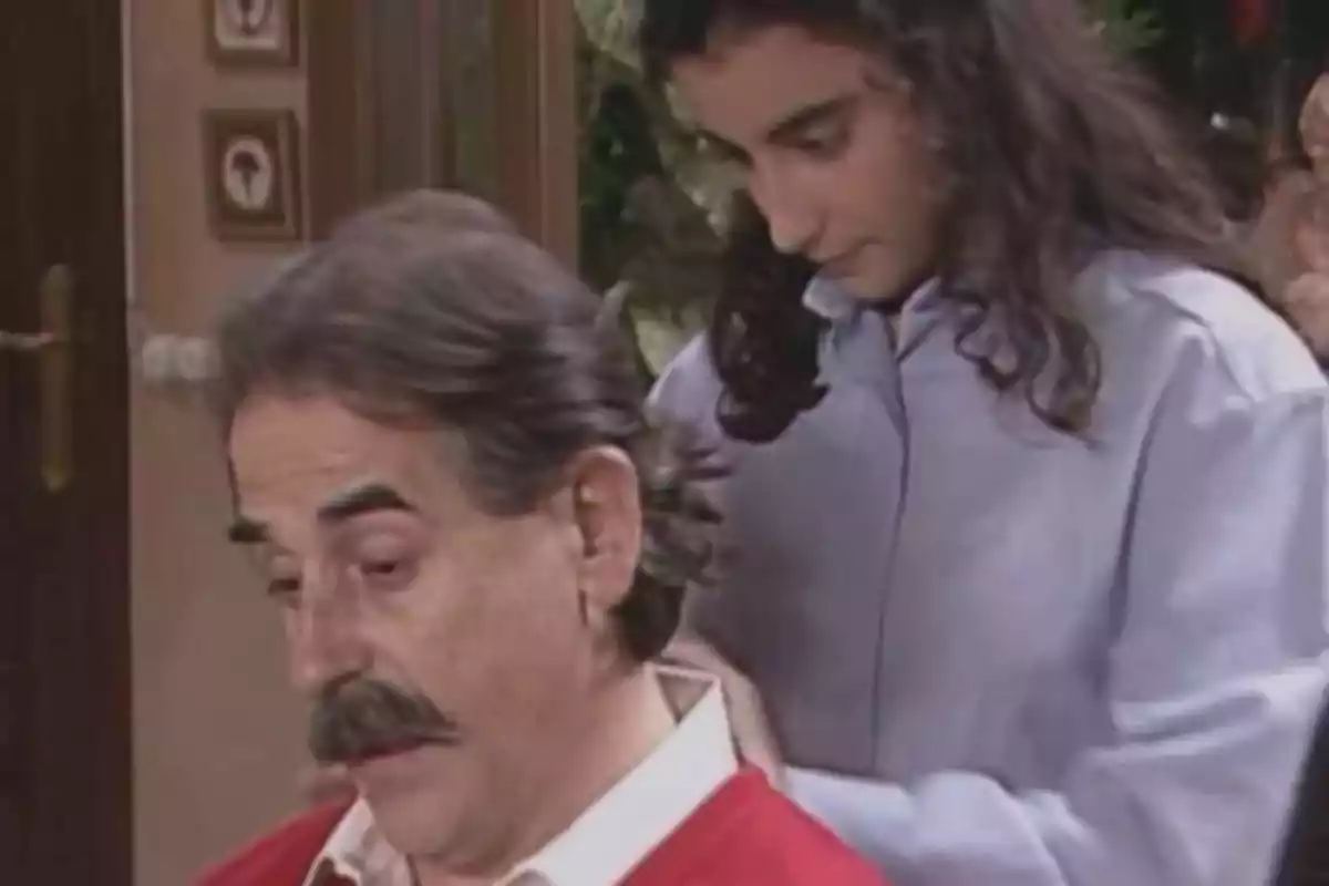 Un hombre de bigote y cabello canoso está sentado mientras una persona joven de cabello largo y oscuro le ayuda a ajustar el cuello de la camisa en una habitación. Un hombre de bigote y cabello canoso está sentado mientras una persona joven de cabello largo y oscuro le ayuda a ajustar el cuello de la camisa en una habitación.