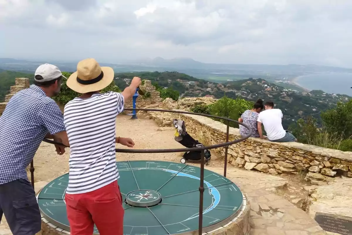 Dues persones observen el paisatge des d’un mirador amb una brúixola gegant al terra mentre una parella seu en un mur de pedra gaudint de la vista panoràmica d’una vall i la costa. Dues persones observen el paisatge des d’un mirador amb una brúixola gegant al terra mentre una parella seu en un mur de pedra gaudint de la vista panoràmica d’una vall i la costa.