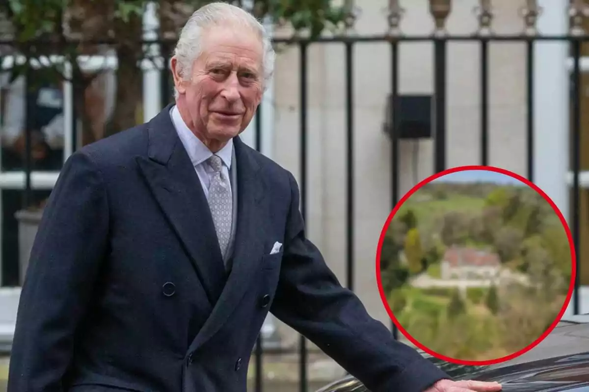 Hombre de cabello canoso con traje oscuro de pie junto a un automóvil, al fondo una cerca de hierro y un círculo rojo que resalta una casa en un paisaje verde desenfocado.
