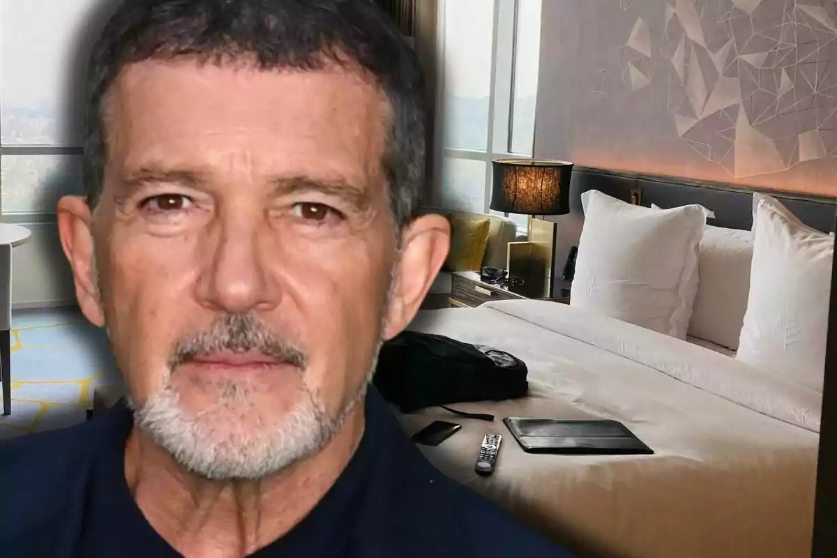 Close-up of Antonio Banderas with a serious expression in front of a modern hotel room with a large bed and personal items on the bed.