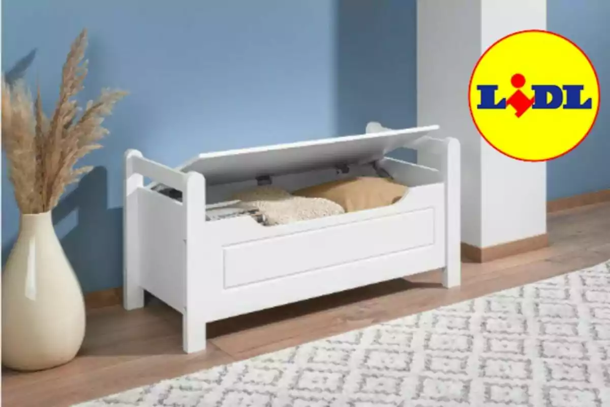 Banco blanco de almacenamiento con cojines y manta en su interior junto a una alfombra y un jarrón decorativo, con el logo de Lidl en la esquina superior derecha. Banco blanco de almacenamiento con cojines y manta en su interior junto a una alfombra y un jarrón decorativo, con el logo de Lidl en la esquina superior derecha.